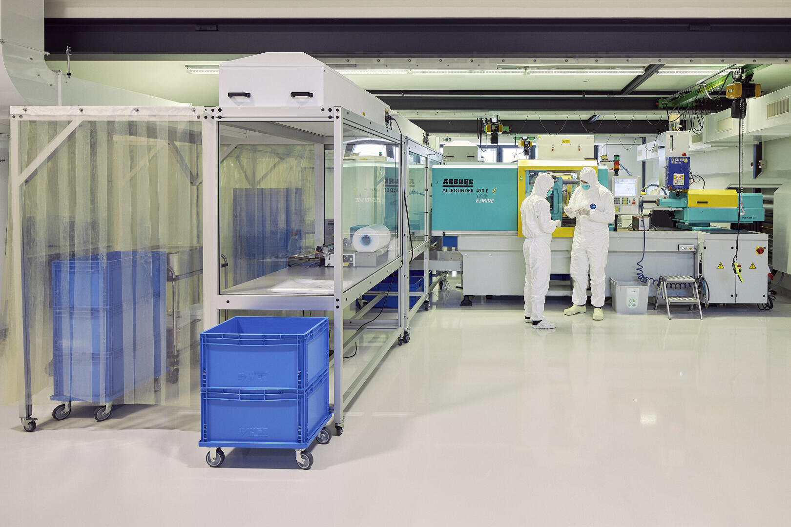 Zwei Personen in weißen Schutzanzügen arbeiten in einem sauberen, modernen Labor mit hellem Licht, großen Maschinen und blauen Plastikbehältern. Die Umgebung wirkt steril und geordnet.