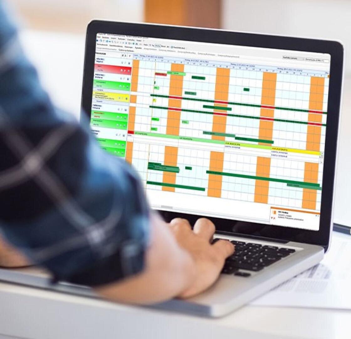 One person operates a laptop on which a colorful planning or project management software with various green, orange and yellow bars is displayed in a grid format.