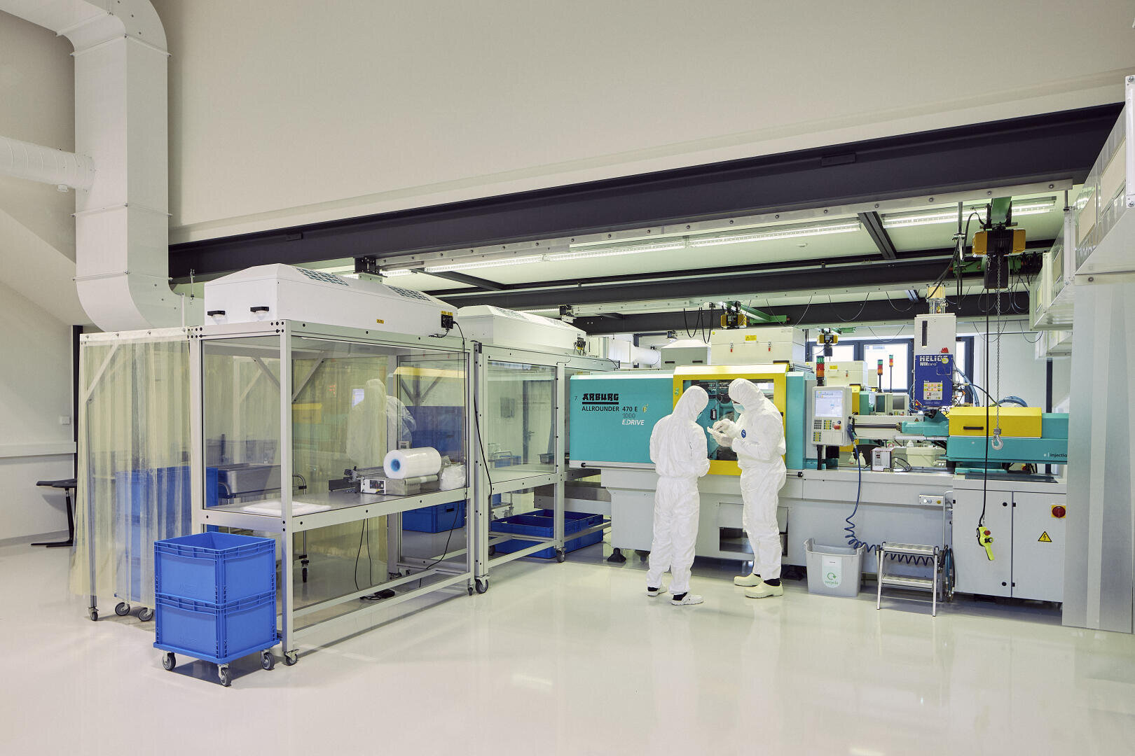 Zwei Personen in Ganzkörperschutzanzügen arbeiten in einem sauberen, modernen Labor mit fortschrittlichen Geräten, einer großen Maschine und Regalen mit blauen Aufbewahrungsbehältern. Die Umgebung ist steril und hell erleuchtet.