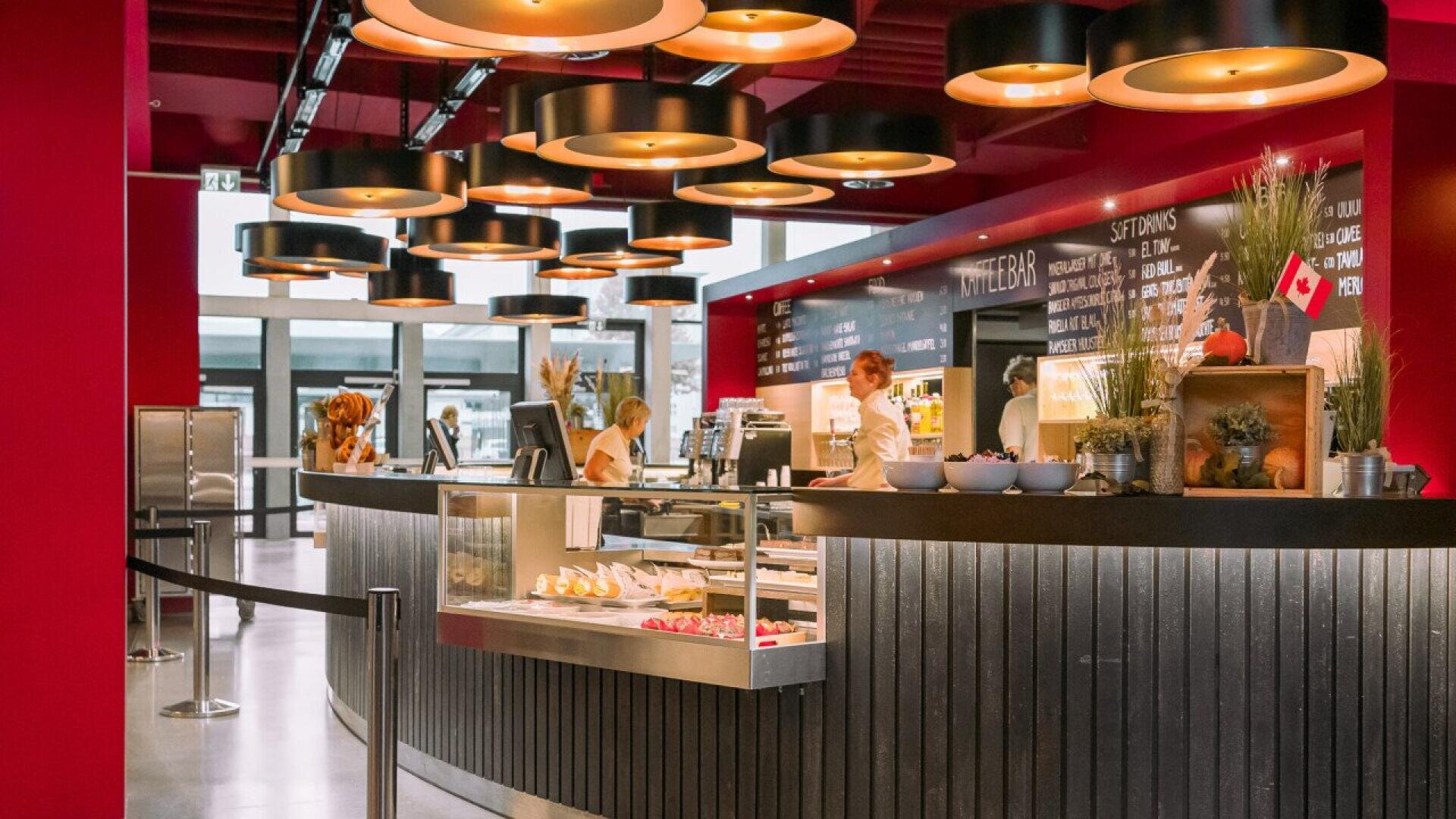 A modern café with red walls, round ceiling lights, and a curved counter displaying pastries. Staff are behind the counter, and a menu board is visible on the wall. Potted plants add decoration to the scene.