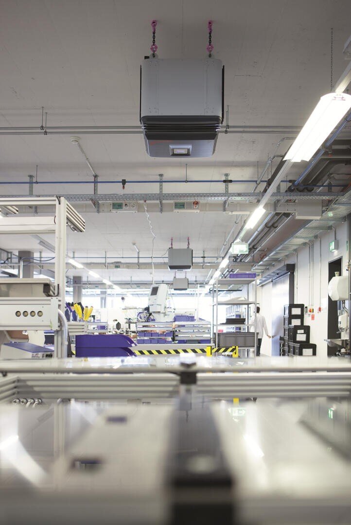 Modern industrial facility with overhead ventilation units, bright lighting, and various machines and equipment arranged in an orderly manner. The environment appears clean and well-organized.