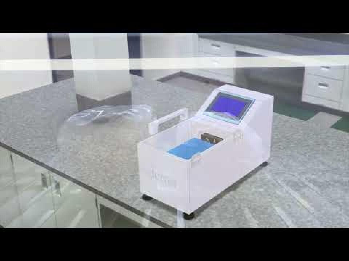 Ein kleines, weißes elektronisches Gerät mit einem Bildschirm steht auf einer Granitarbeitsplatte in einer Laborumgebung. In der Nähe befindet sich eine durchsichtige Plastikschutzbrille.