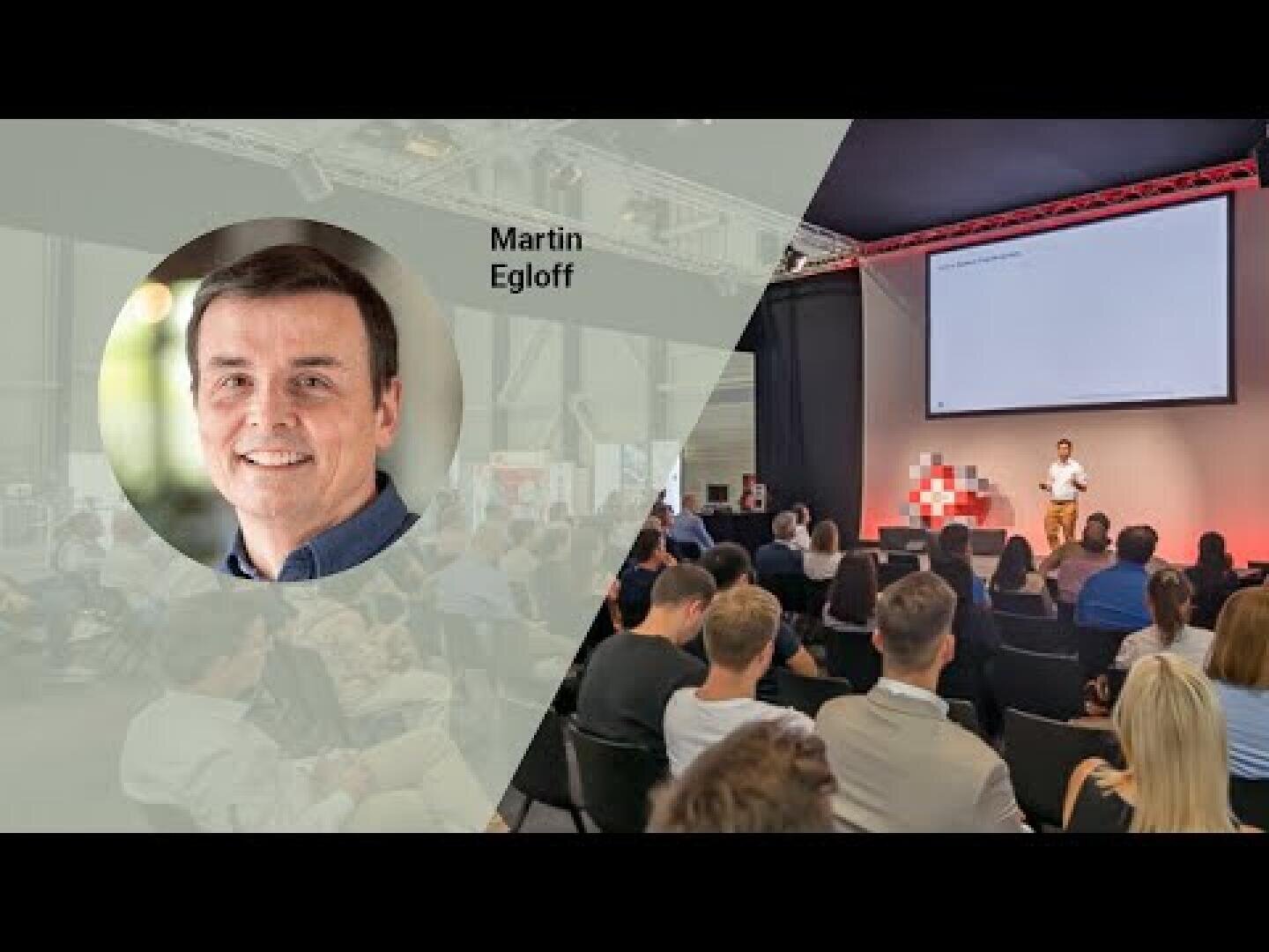 Ein großer Raum mit Menschen, die vor einer Bühne sitzen, auf der ein Redner vorträgt; ein kreisförmiger Einschub zeigt das Porträt eines lächelnden Mannes mit der Aufschrift Martin Egloff.