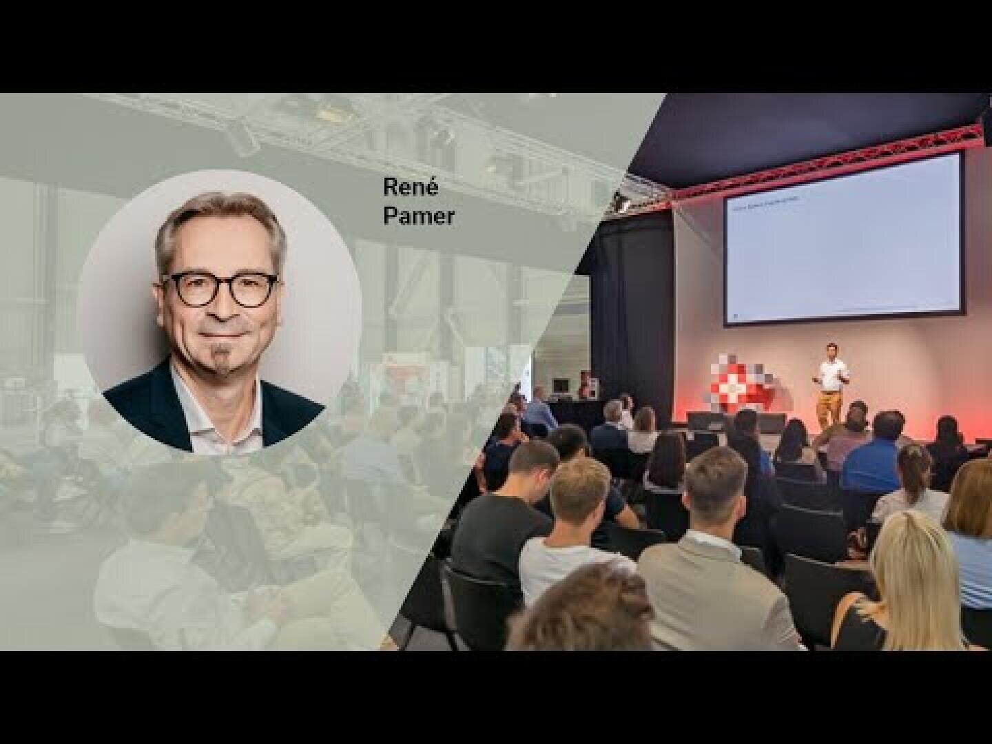 Ein Mann spricht auf der Bühne zu einem Publikum in einem Konferenzraum. Auf der linken Seite ist das Foto eines Mannes mit Brille eingeblendet, das die Bezeichnung René Pamer trägt. Die Umgebung ist professionell, mit einem großen Bildschirm und roter Beleuchtung.