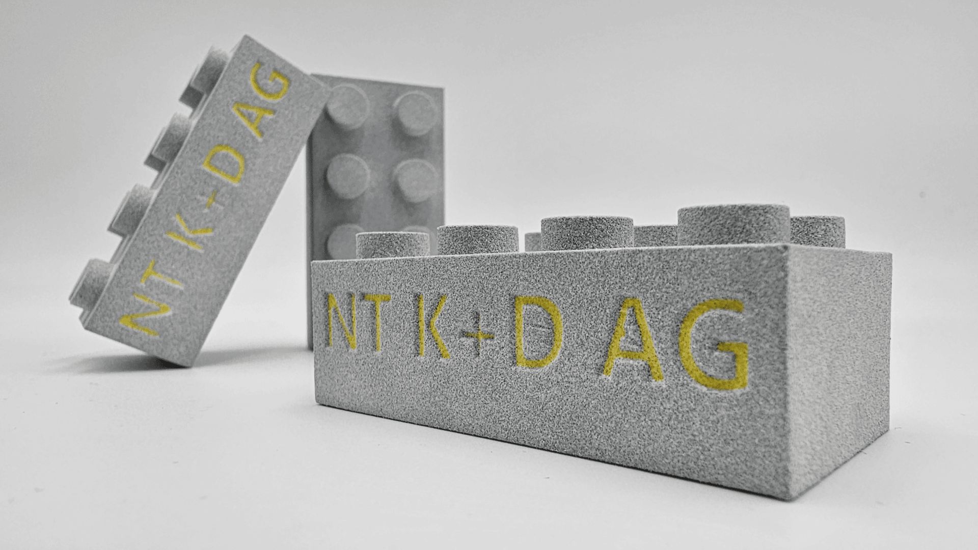 Drei graue Spielzeugbausteine mit strukturierter Oberfläche sind auf weißem Untergrund angeordnet. Auf jedem Baustein steht an der Seite gelb der Text „NT K-DAG“.