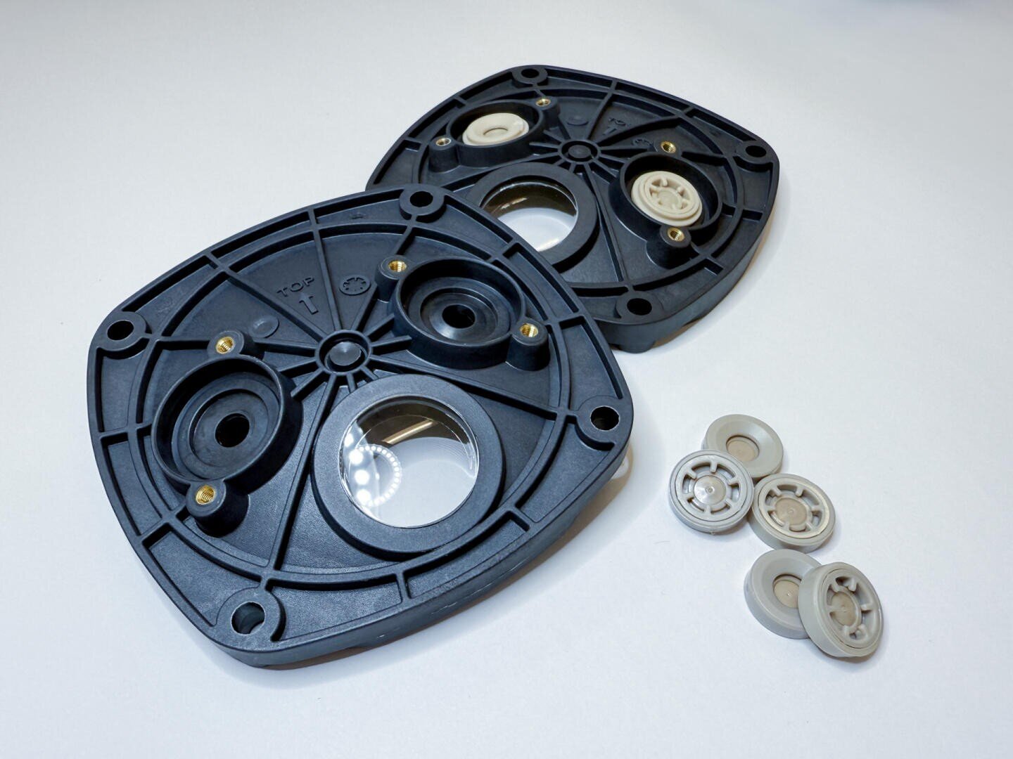 Two black mechanical plastic parts with circular openings and several screw holes are located on a white surface. Four small beige-colored, gear-like components are located next to the parts.