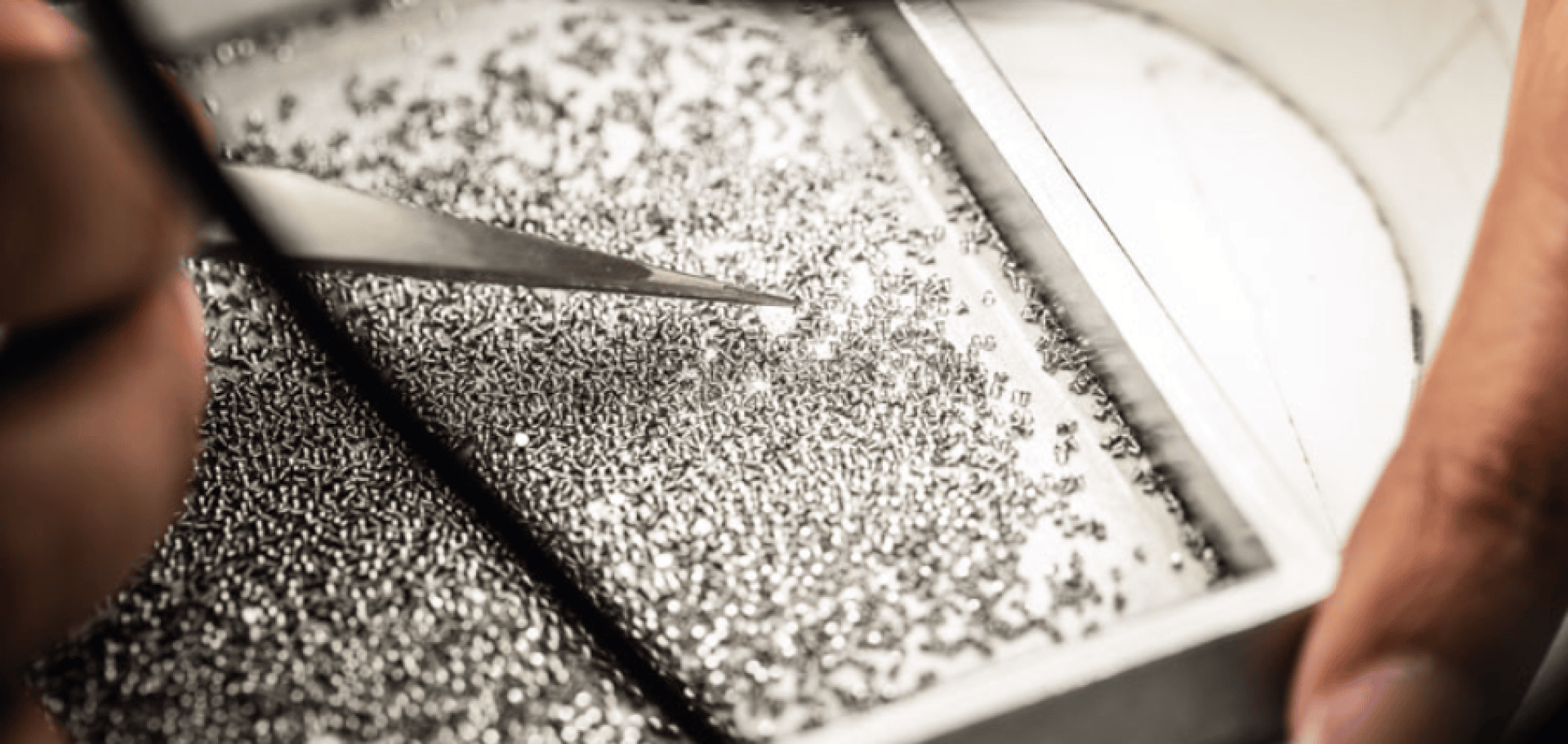 Close-up of hands using tweezers to pick up small, shiny metal pieces from a tray, possibly in a precision manufacturing or sorting process.