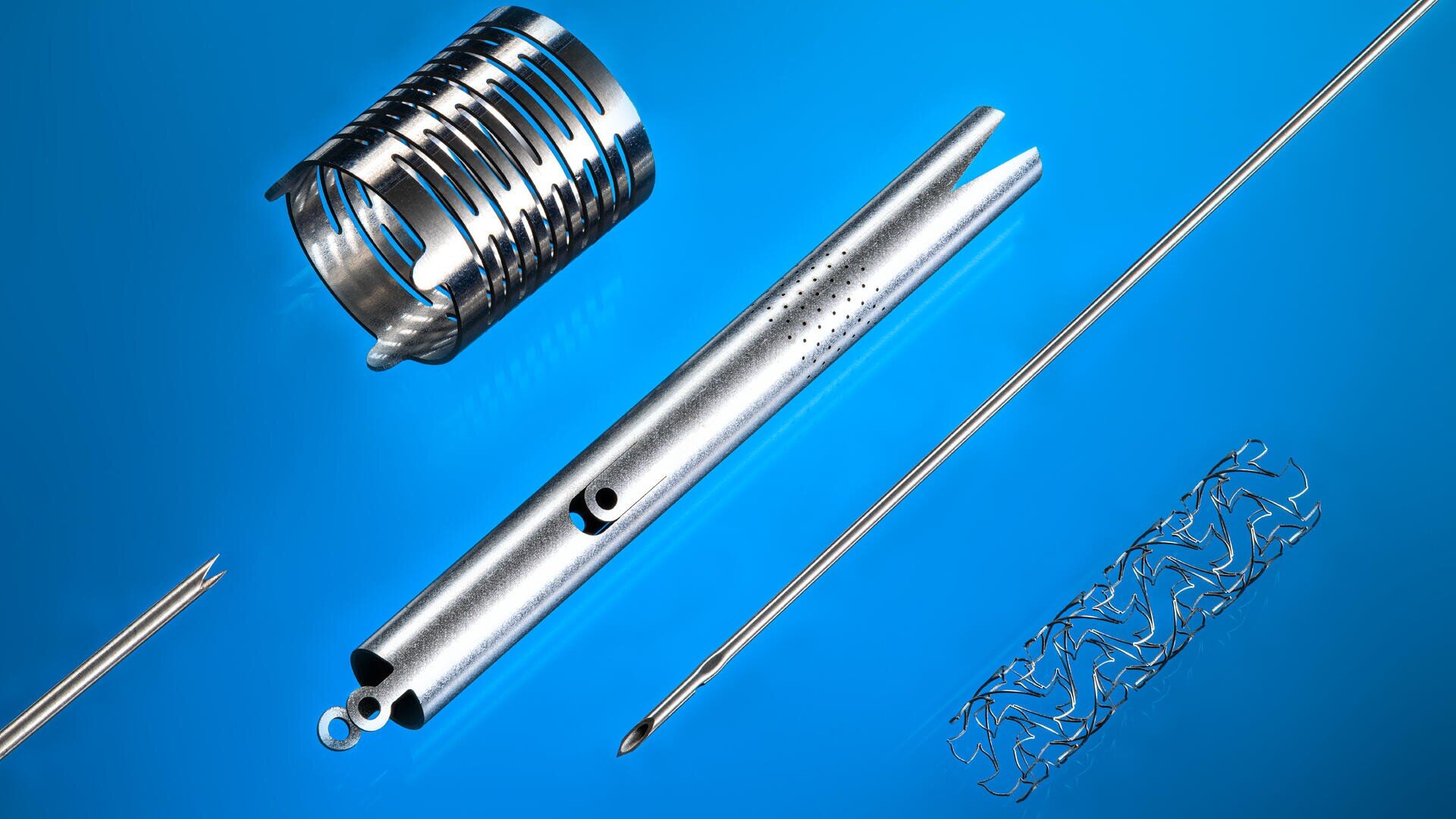 Auf einem blauen Hintergrund sind verschiedene metallisch glänzende medizinische Geräte angeordnet, darunter ein Rohr mit Löchern, eine Spule, zwei spitze Nadeln und ein Netzstent.