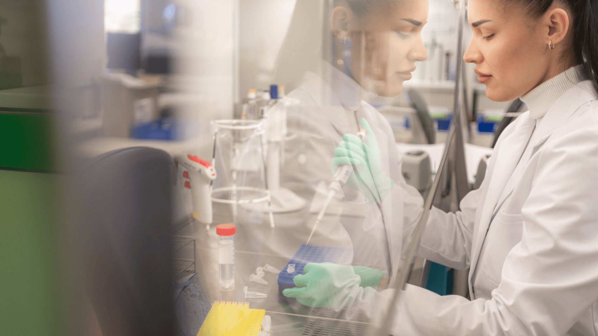 Eine Wissenschaftlerin in einem weißen Laborkittel und mit grünen Handschuhen benutzt eine Pipette in einem Labor, wobei ihr Spiegelbild auf einer Glasscheibe neben ihr zu sehen ist. Auf dem Arbeitstisch sind Laborgeräte und Zubehör zu sehen.