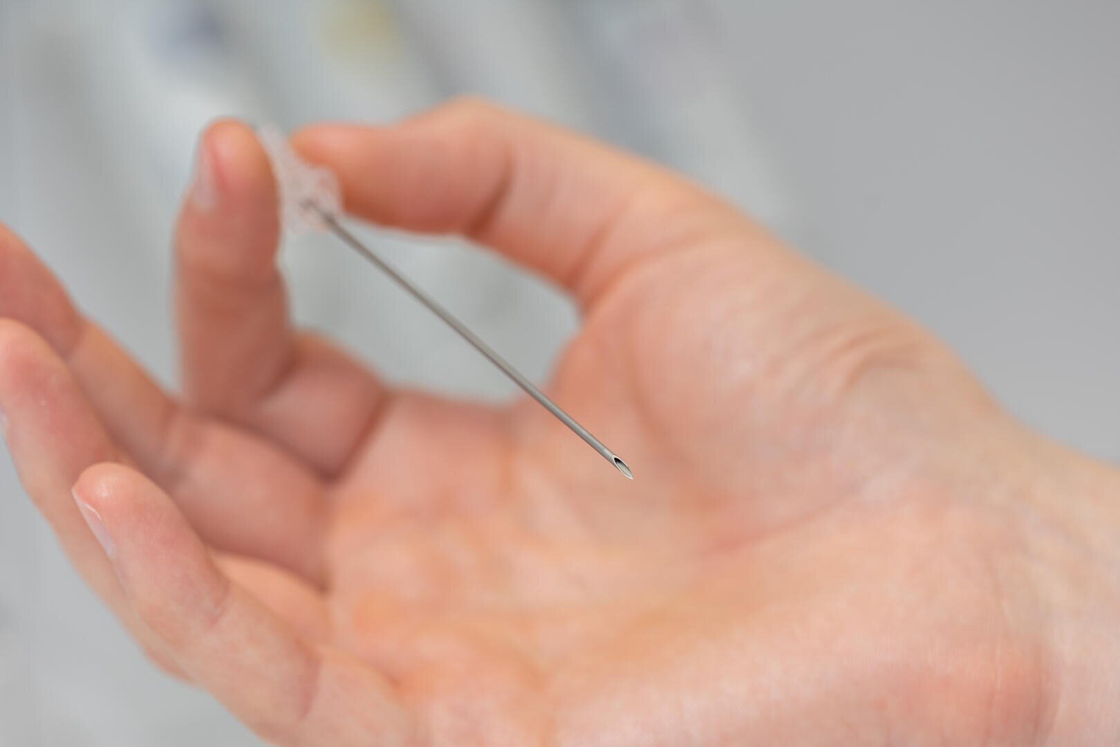 Close-up of a person holding a thin medical needle, with the sharp tip pointing towards the camera and the background out of focus.