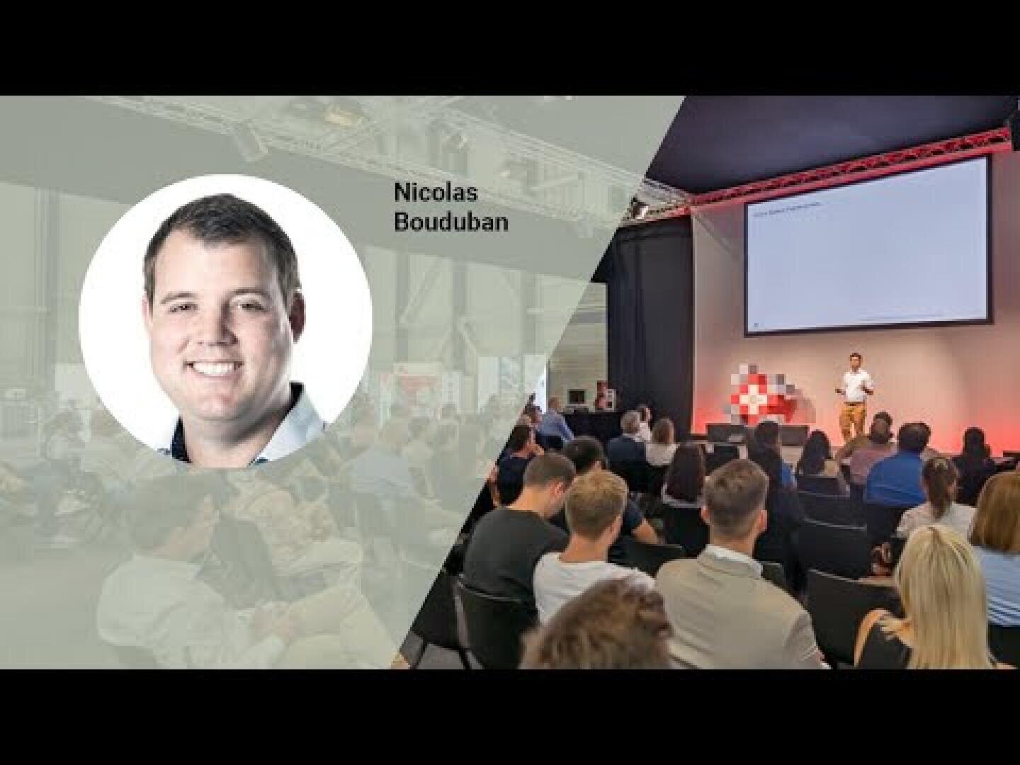 Ein Mann spricht auf der Bühne vor einem sitzenden Publikum bei einer Konferenz. Eine Einblendung zeigt ein Kopfbild von Nicolas Bouduban mit seinem Namen daneben. Hinter dem Redner ist ein großer Bildschirm zu sehen.