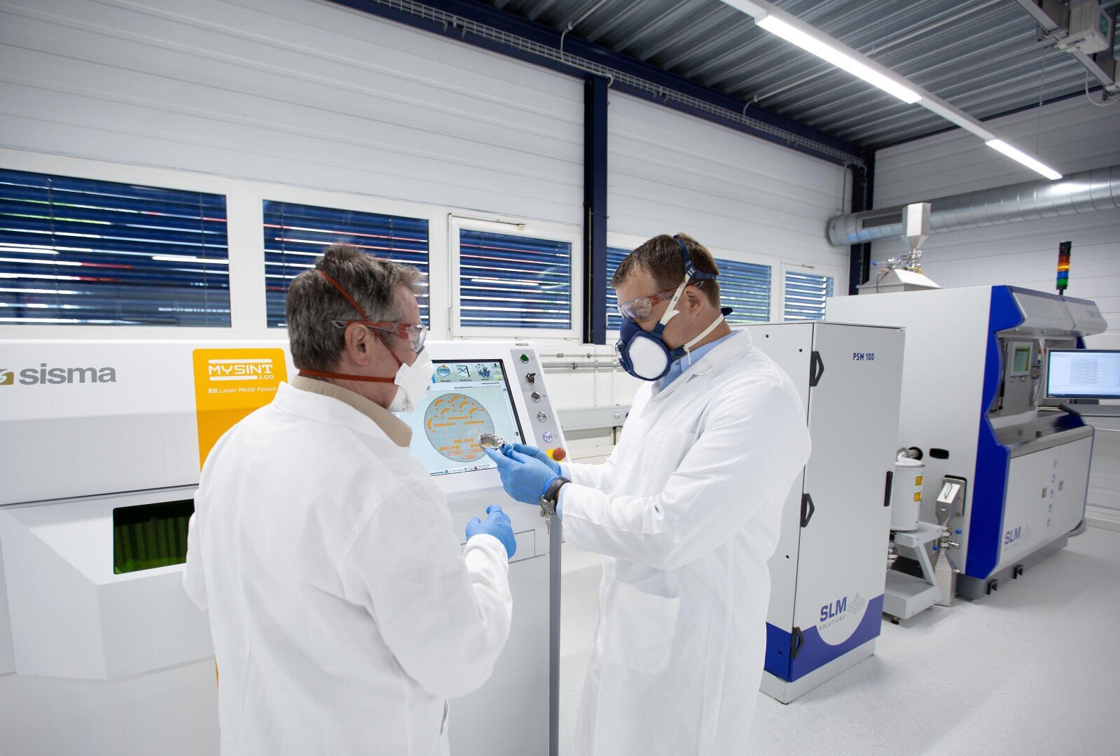 Zwei Personen in Laborkitteln und Schutzkleidung bedienen eine Touchscreen-Maschine in einem modernen Labor mit modernster Ausstattung und weißen Wänden.