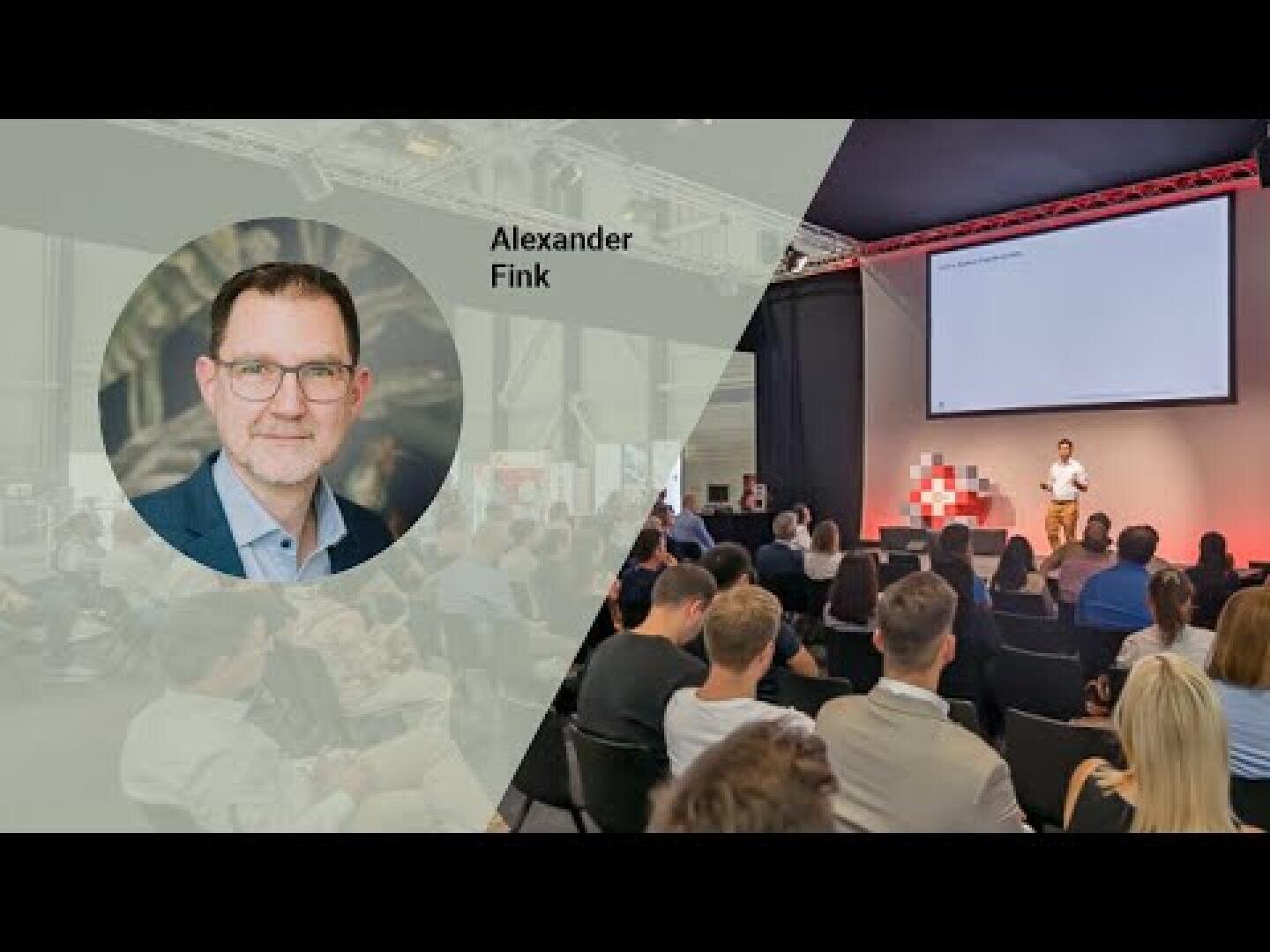 A presentation event with an audience facing a stage where a person is speaking near a large screen. An inset shows a portrait of a man labeled Alexander Fink.