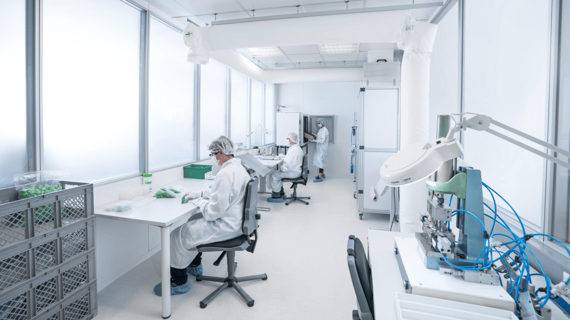 Three people wearing lab coats, hairnets, and gloves work at desks in a bright, modern laboratory with large windows, scientific equipment, and storage bins.