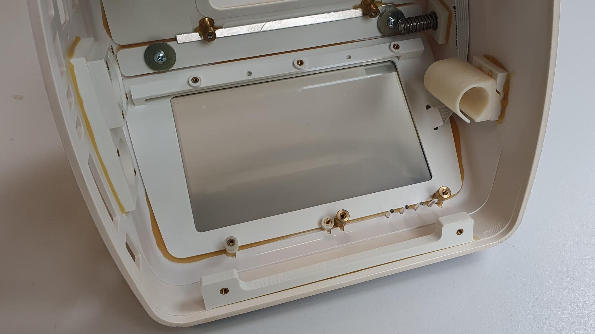 Close-up of the inside of a white plastic device showing a rectangular transparent plate, several screws, metal parts and a spring mechanism on top resting on a white surface.