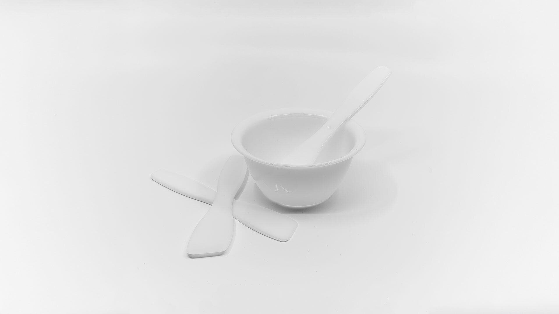 Eine weiße Plastikschüssel mit einem darin liegenden Spatel und zwei weiteren Spateln, die flach neben der Schüssel liegen, alles vor einem schlichten weißen Hintergrund.