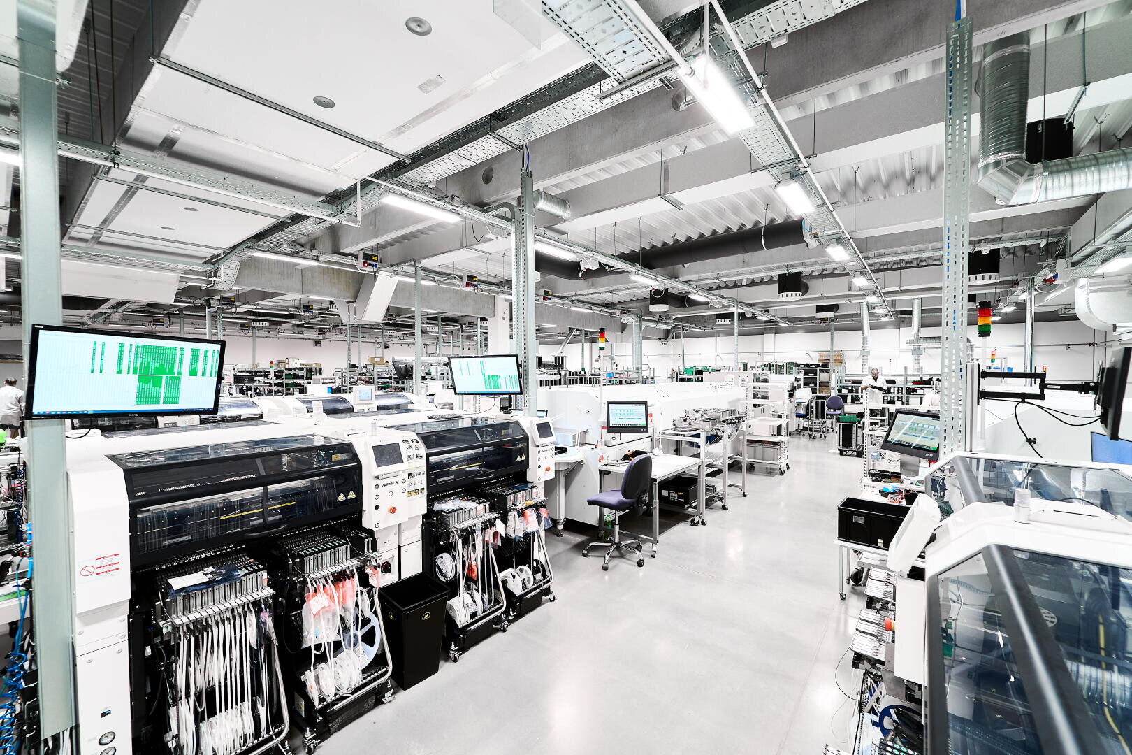 A bright, modern electronics manufacturing facility with automated machinery, computer monitors, workstations, and organized components on a polished concrete floor under exposed ceilings.