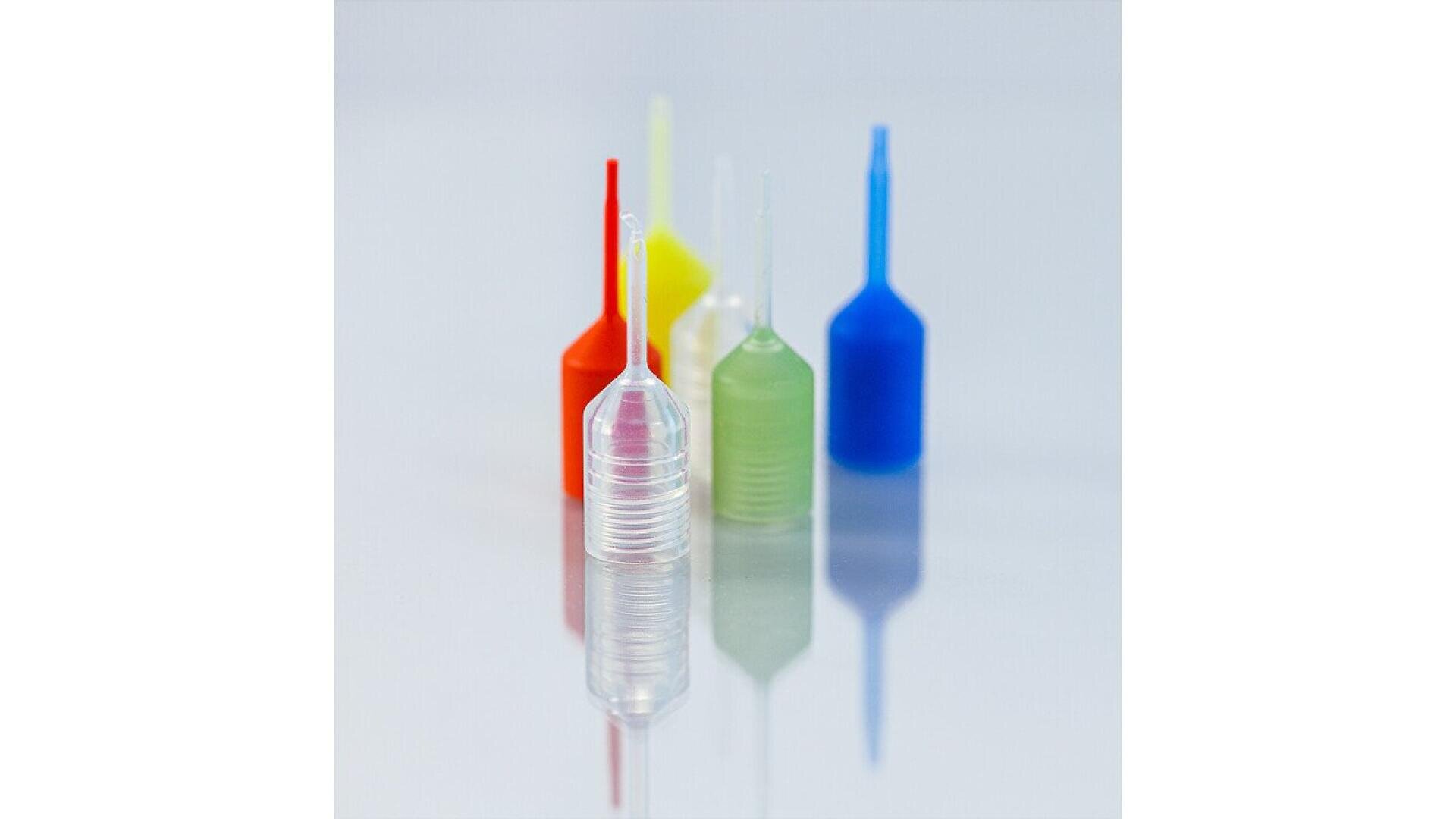 Five small plastic squeeze bottles with long, narrow nozzles, each in different translucent colors (clear, red, yellow, green, and blue), are arranged on a reflective surface against a light background.