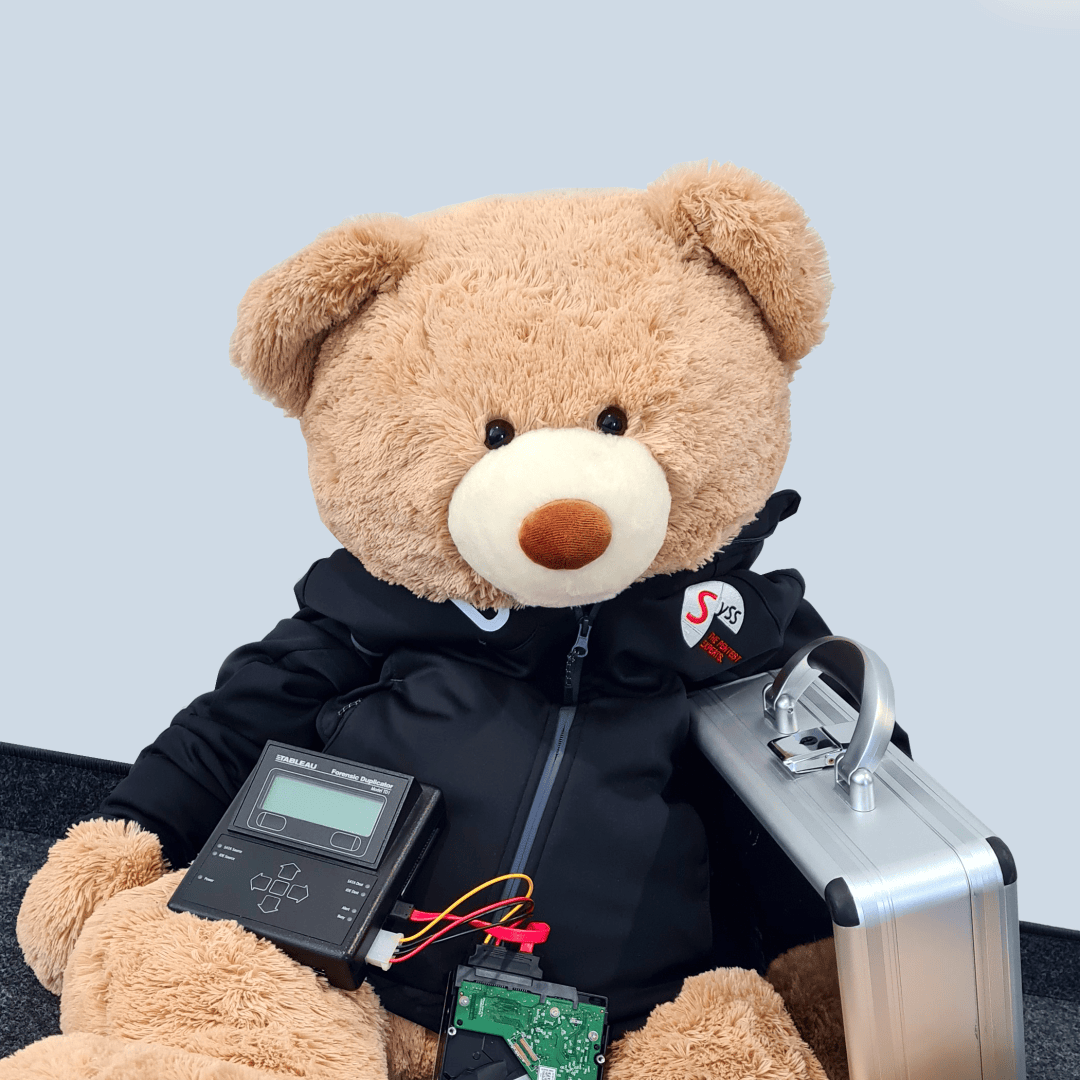 Ein großer brauner Teddybär in einer schwarzen Jacke sitzt auf dem Boden und hält ein silbernes Gehäuse, ein elektronisches Gerät mit Drähten und eine grüne Platine vor einem schlichten hellen Hintergrund.
