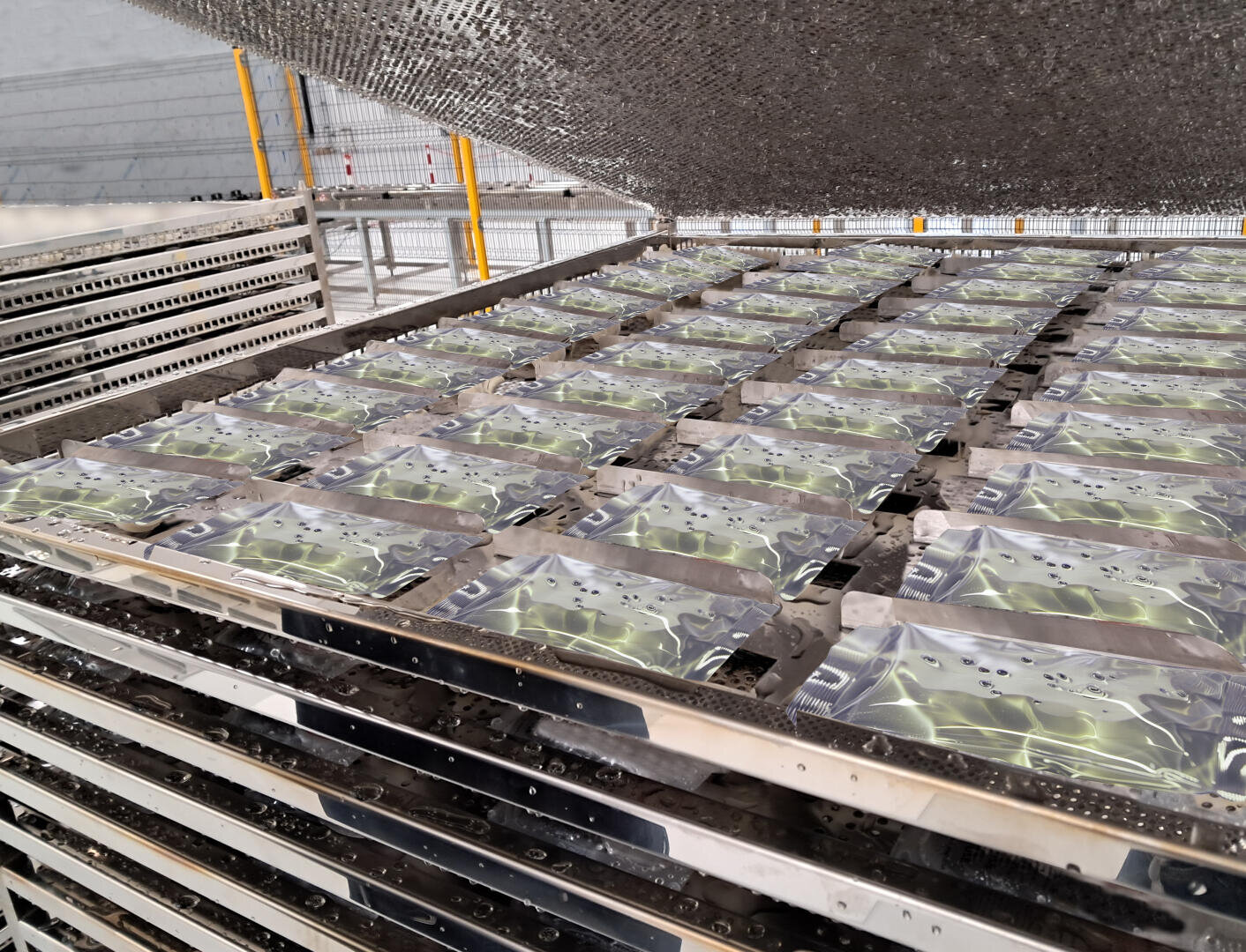 Stapel von rechteckigen Metallschalen, die mit einer klaren Flüssigkeit und grünlichen Substanzen, möglicherweise Algen oder Laborproben, gefüllt sind, sind in einem Gittermuster auf Metallgestellen in einer Industrie- oder Forschungseinrichtung angeordnet.