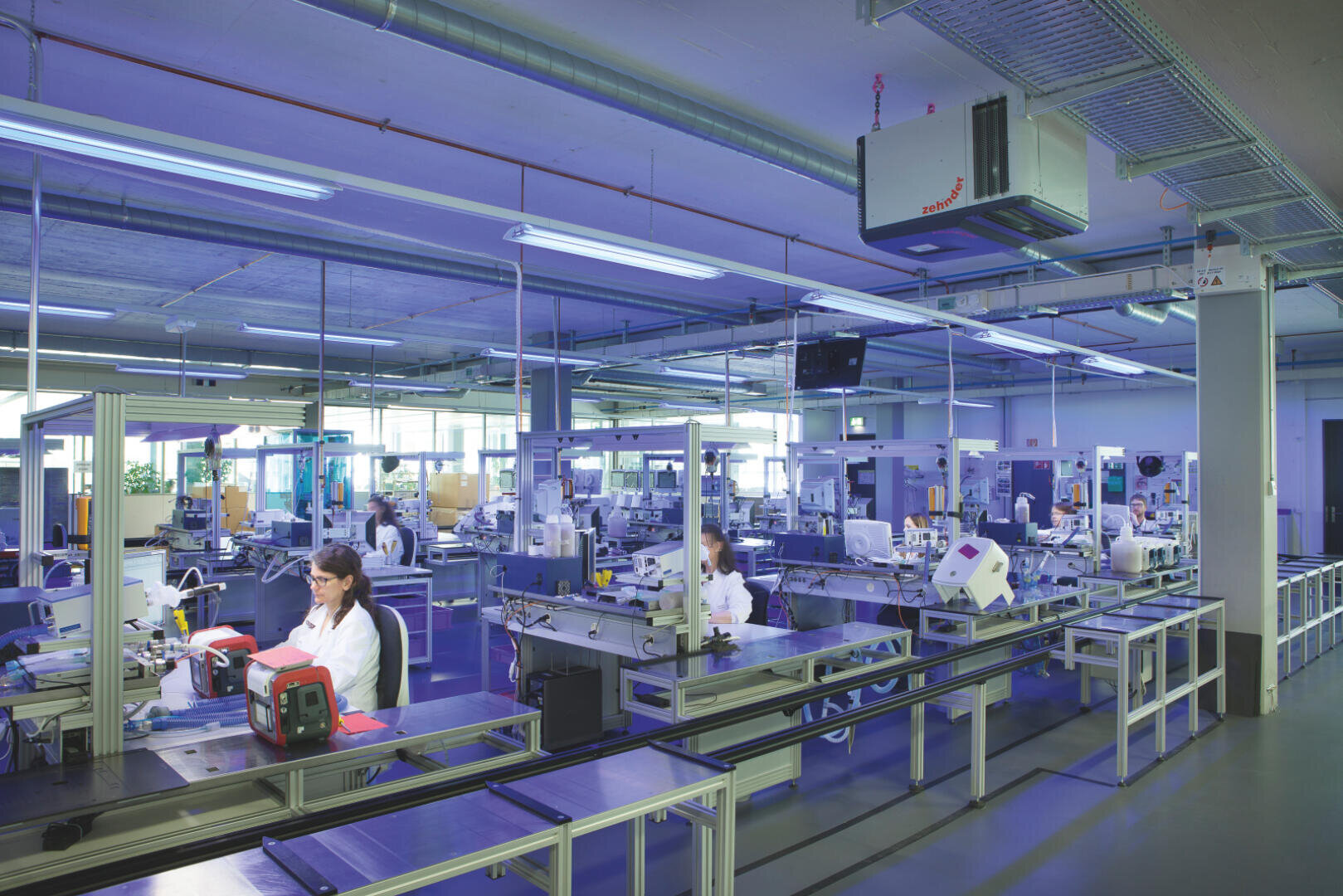 A modern, brightly lit factory laboratory with workers in white lab coats operating machines and assembling products at organized workstations under blue light.