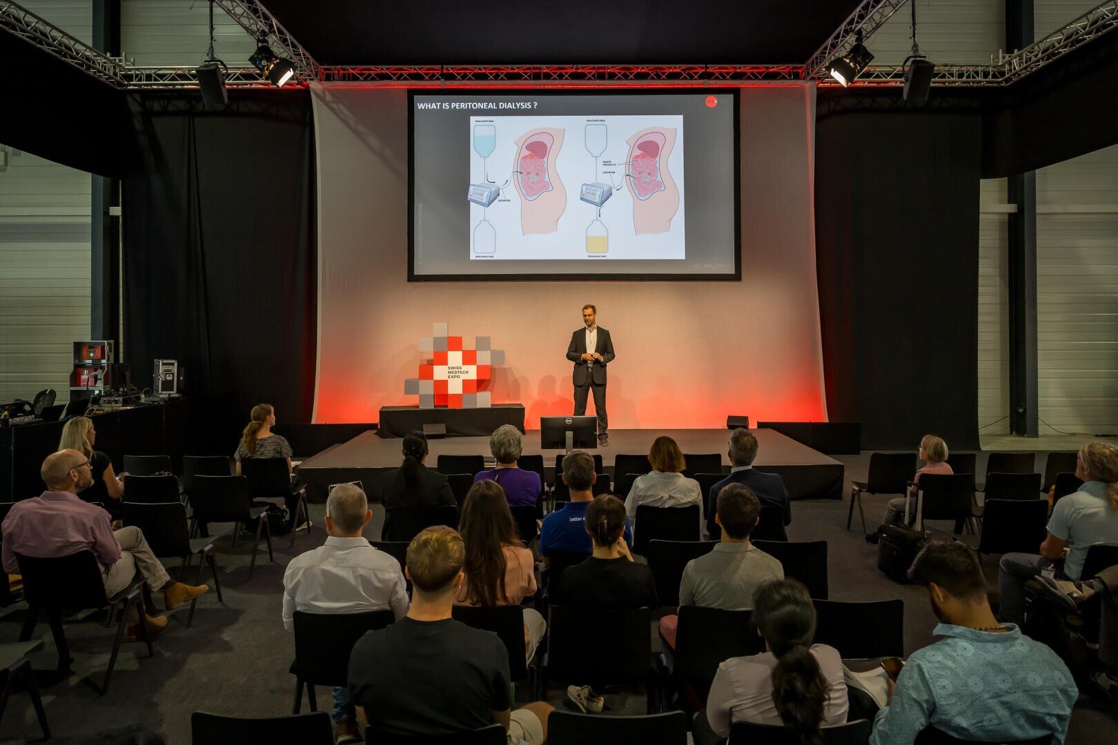 Ein Redner im Anzug präsentiert den Zuhörern in einem modernen Konferenzraum mit roter Beleuchtung und einem karierten Bühnenhintergrund medizinische Diagramme auf einem großen Bildschirm.