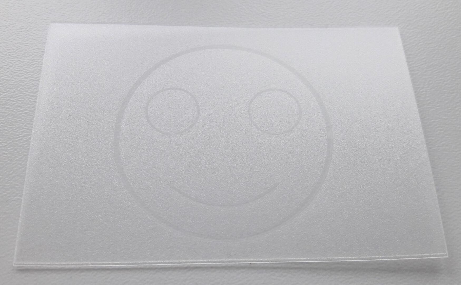 Eine mattierte, durchscheinende Plastikfolie mit einem leicht geprägten Smiley-Gesicht mit zwei Augen und einem lächelnden Mund auf einer strukturierten weißen Oberfläche.