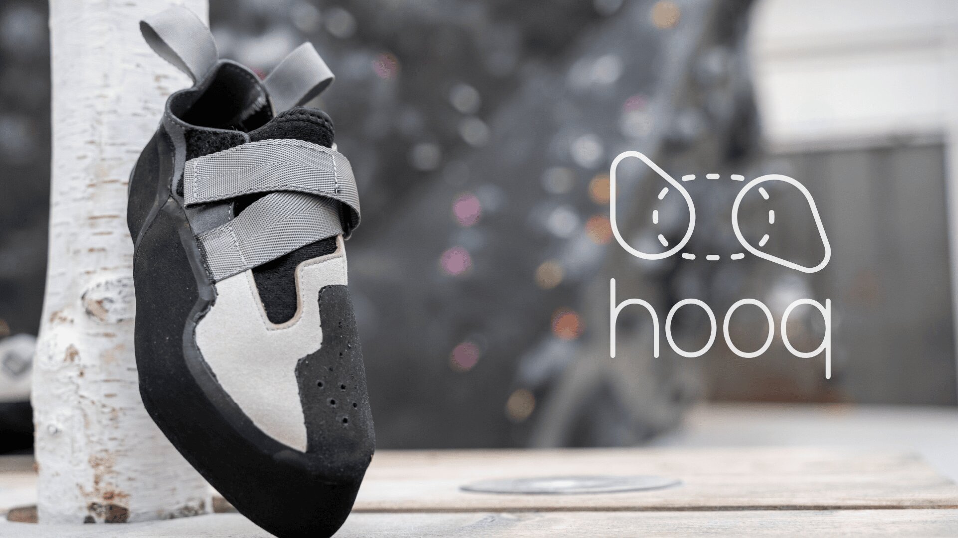 Ein Kletterschuh in grauer und schwarzer Farbe hängt an einem Holzpfosten, auf dem rechts der Markenname hooq und ein Logo abgebildet sind. Im Hintergrund ist eine unscharfe Kletterwand mit farbigen Griffen zu sehen.