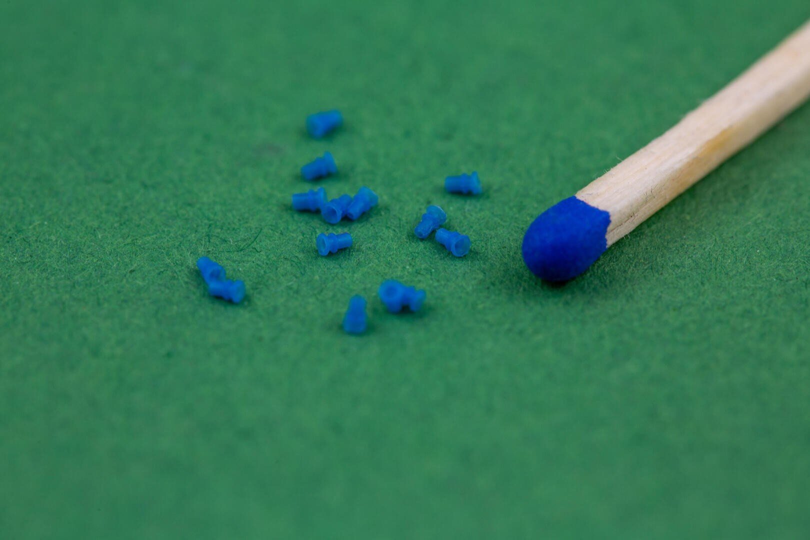 Nahaufnahme eines Streichholzes mit einer blauen Spitze neben mehreren winzigen blauen Objekten, die auf einer grünen Fläche verstreut sind. Die Objekte erscheinen sehr klein im Vergleich zu dem Streichholz.