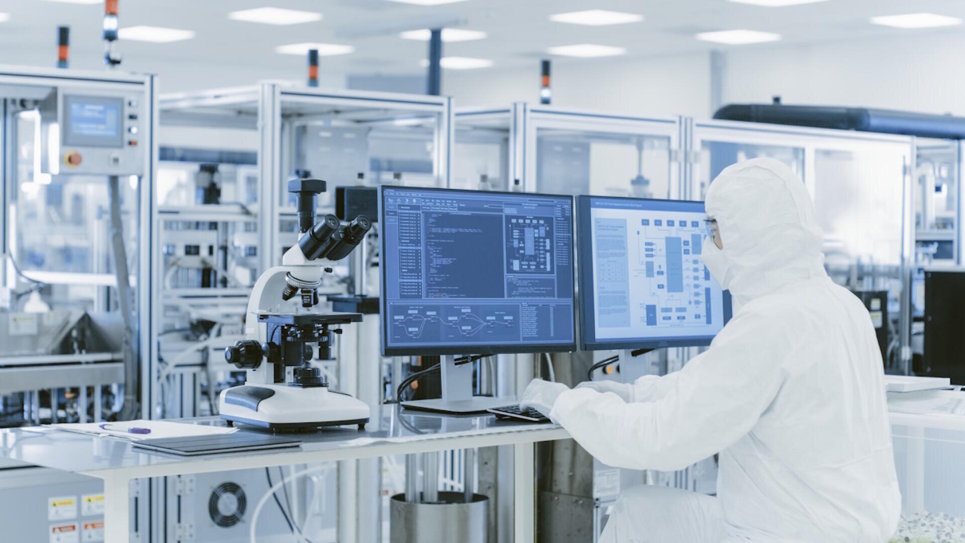 A person in full protective clothing is working at a computer in a high-tech laboratory, with a microscope and laboratory equipment on his desk and technical data displayed on large monitors.