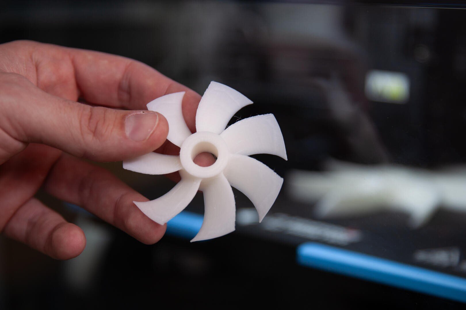 Eine Hand, die einen weißen, 3D-gedruckten Ventilator oder eine Turbinenschaufel hält, mit einem unscharfen Hintergrund, der auf einen 3D-Drucker im Einsatz schließen lässt.