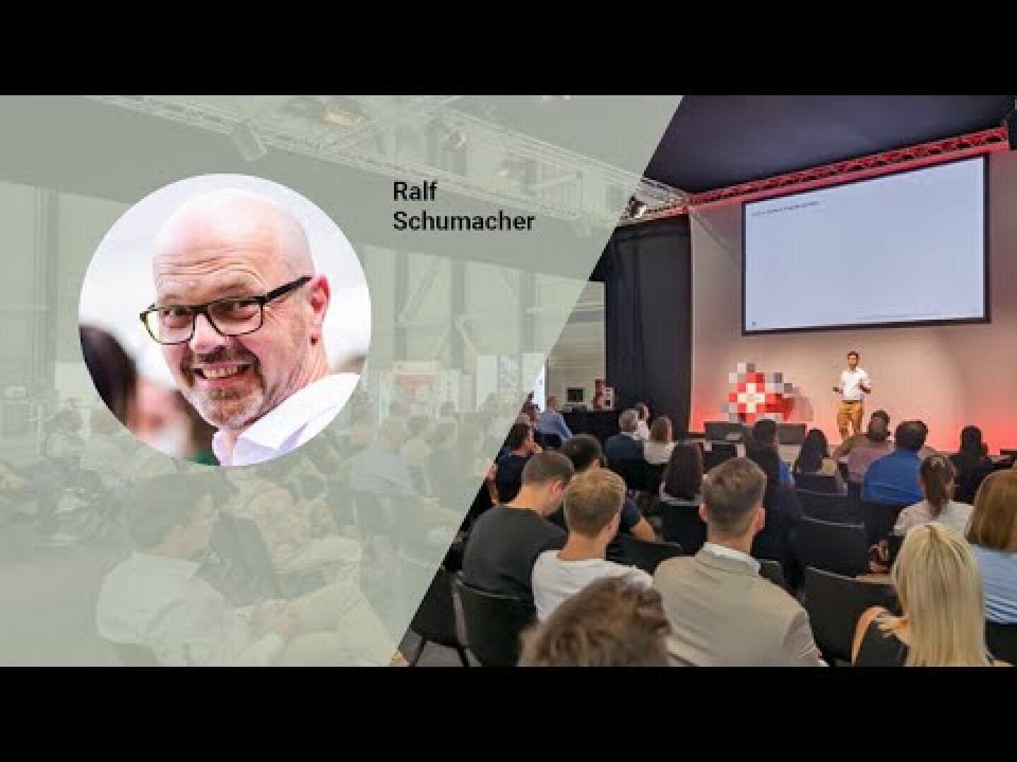Ein Redner steht auf der Bühne und hält einen Vortrag vor einem Publikum in einem modernen Veranstaltungsraum. Auf der linken Seite zeigt ein kreisförmiger Einschub einen lächelnden Mann mit der Aufschrift Ralf Schumacher. Das Publikum ist der Bühne zugewandt und hört aufmerksam zu.