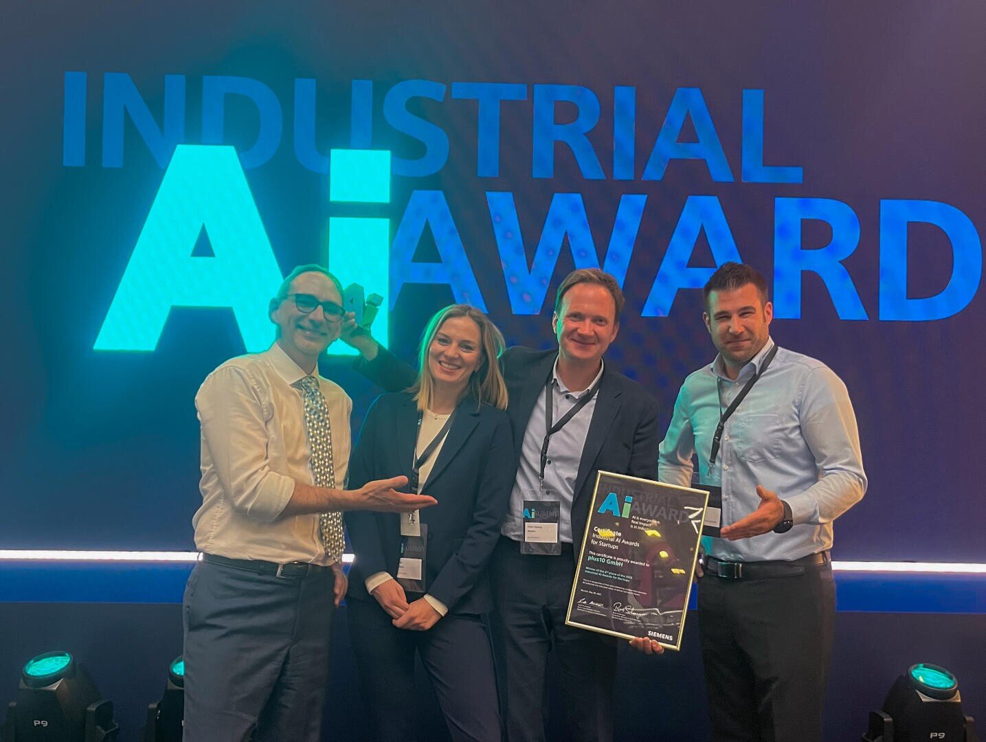 Vier Personen in Geschäftskleidung stehen lächelnd vor einem großen Schild mit dem Industrial AI Award, halten eine gerahmte Urkunde in der Hand und zeigen mit stolzen Gesten, dass sie eine Auszeichnung erhalten haben.