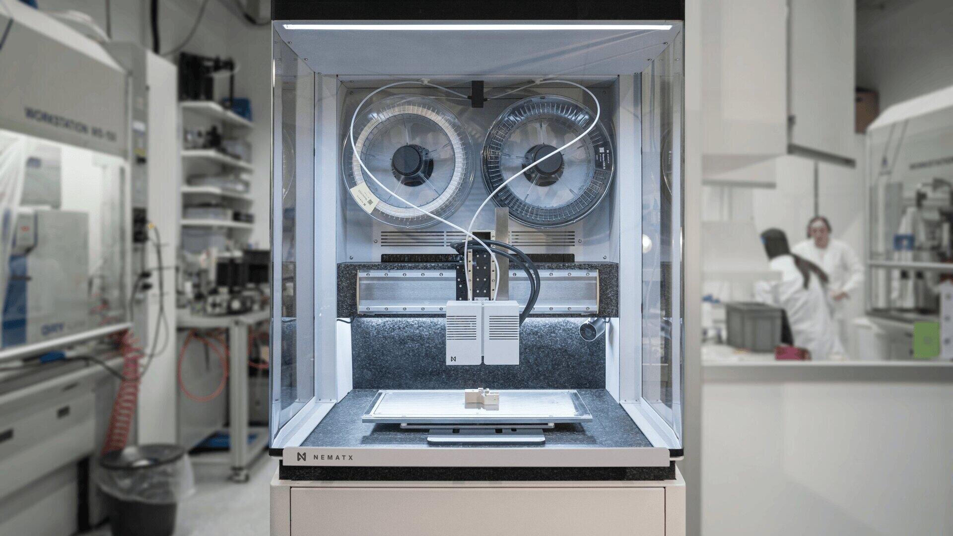 Ein 3D-Biodrucker mit transparentem Gehäuse steht in einem modernen Labor, mit zwei großen Rollen und einem Präzisionsdruckkopf; im Hintergrund sind Laborgeräte und Menschen in weißen Kitteln zu sehen.
