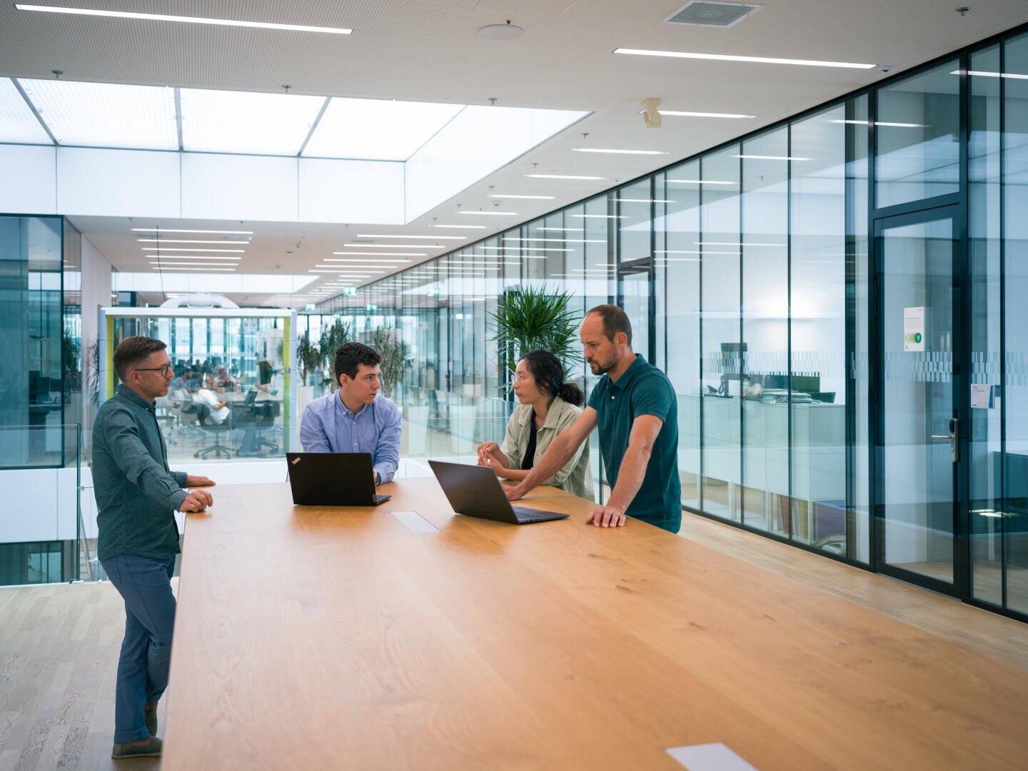 Vier Personen stehen um einen langen Holztisch in einem modernen Büro und besprechen ihre Arbeit mit geöffneten Laptops. In der hellen, geräumigen Umgebung sind große Glaswände und Zimmerpflanzen zu sehen.