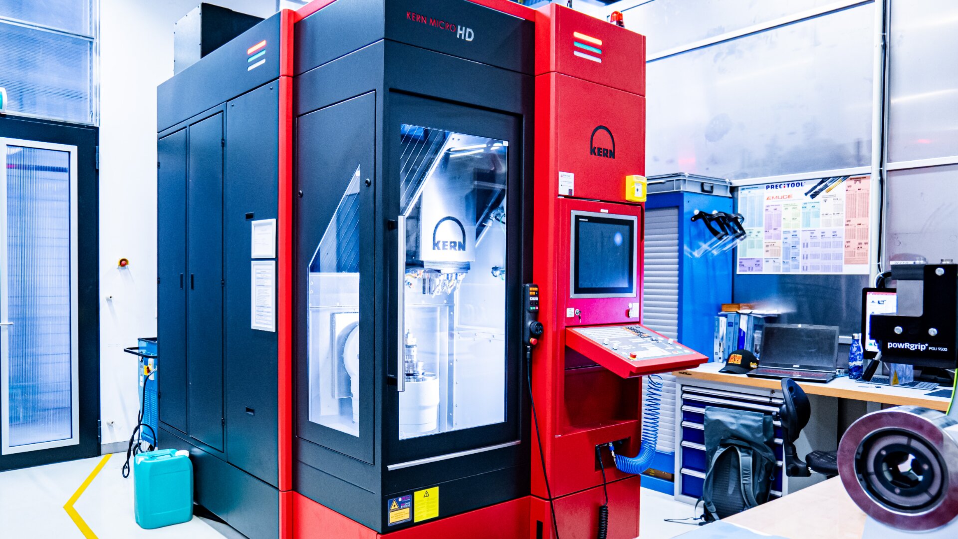 Eine große rot-schwarze CNC-Industriemaschine mit der Aufschrift KERN MICRO HD steht in einem hellen, modernen Labor mit einem Computerbildschirm, einem Arbeitsplatz, Werkzeugen und technischen Tabellen an der Wand daneben.