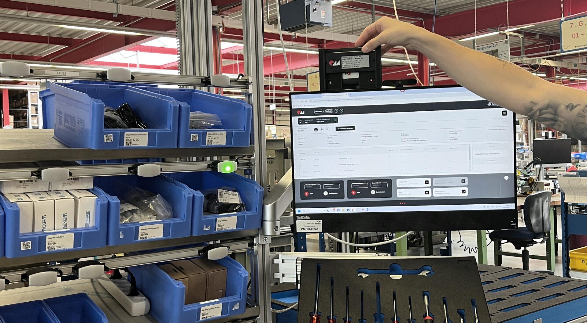 Eine Person hält ein kleines Gerät in der Nähe eines Monitors, auf dem eine Bestandsverwaltungssoftware in einer Fabrik angezeigt wird. Blaue Behälter mit beschrifteten Teilen und Werkzeugen sind in Regalen in der Nähe aufgereiht. Die Umgebung wirkt industriell und geordnet.
