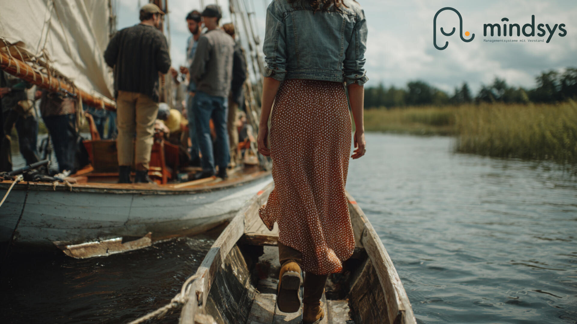 A woman in a red patterned skirt walks along a narrow wooden walkway towards a group of people gathered on a sailing boat, surrounded by reeds and water. The "mindsys" logo appears in the top right-hand corner.
