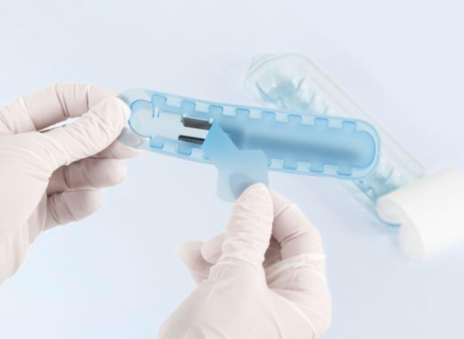 A person wearing white gloves holds a light blue plastic dental tool, possibly a floss pick, with a clear plastic case in the background on a light blue surface.