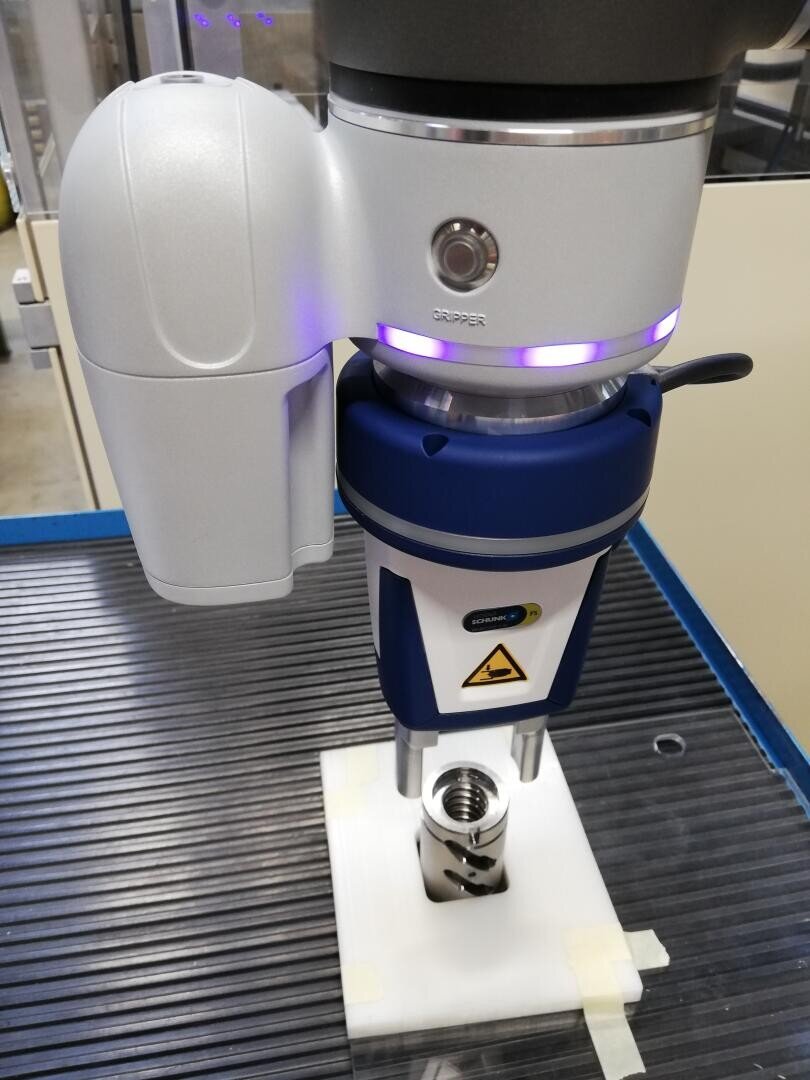 Close-up of an industrial robotic arm with a gripping device positioned over a metal part on a white platform, probably for automated assembly or testing.