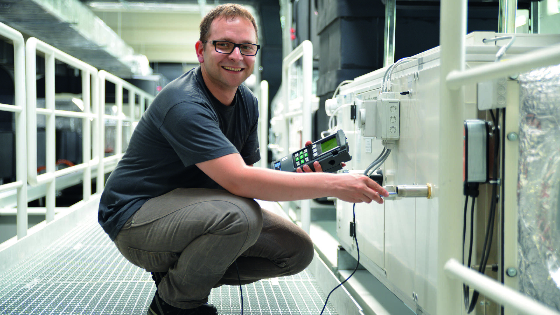 Eine Person mit Brille und dunklem T-Shirt hockt lächelnd auf einem Metallsteg, während sie in einer Industrie- oder Laborumgebung ein elektronisches Gerät zur Überprüfung oder Kalibrierung von Ausrüstung verwendet.