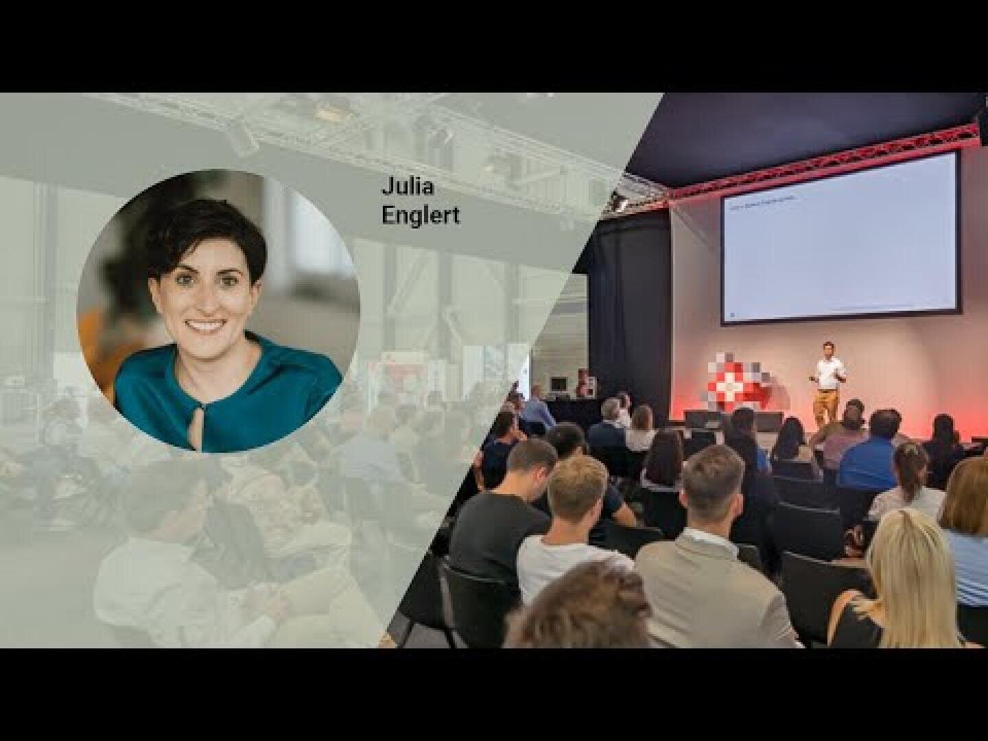 Ein Foto von einer Konferenz mit einem Publikum, das einem Redner auf der Bühne gegenübersteht. Auf der linken Seite ist ein rundes Porträt von Julia Englert zu sehen, daneben steht ihr Name. Im Hintergrund sind die Bühne und das Publikum zu sehen.