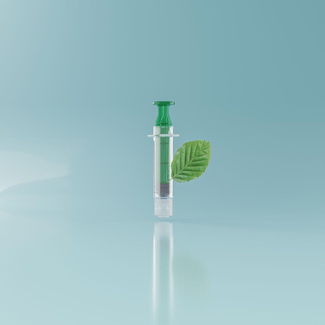 A syringe with a green plunger and a green liquid inside stands upright on a light blue background. A single green leaf is attached to the side of the syringe, symbolizing an eco-friendly or natural concept.