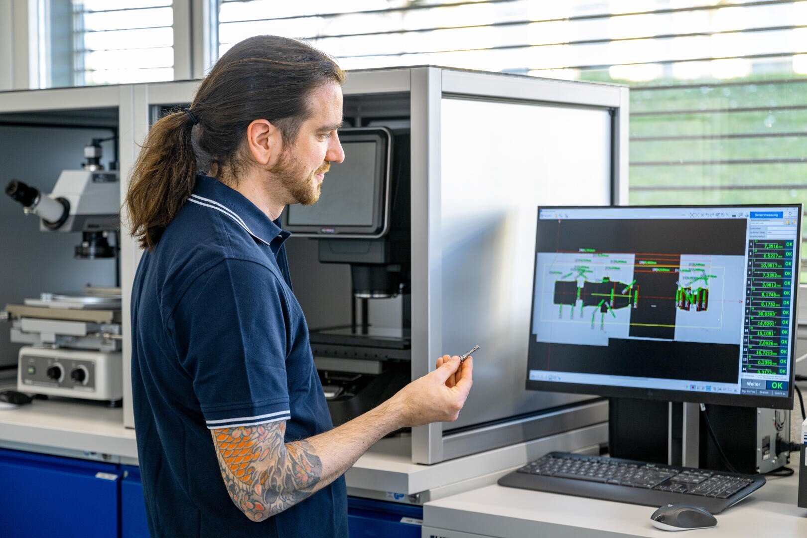 A man with a ponytail and tattooed arm stands in a lab, holding a small object and looking at a computer monitor displaying technical diagrams and data. Laboratory equipment and microscopes are in the background.