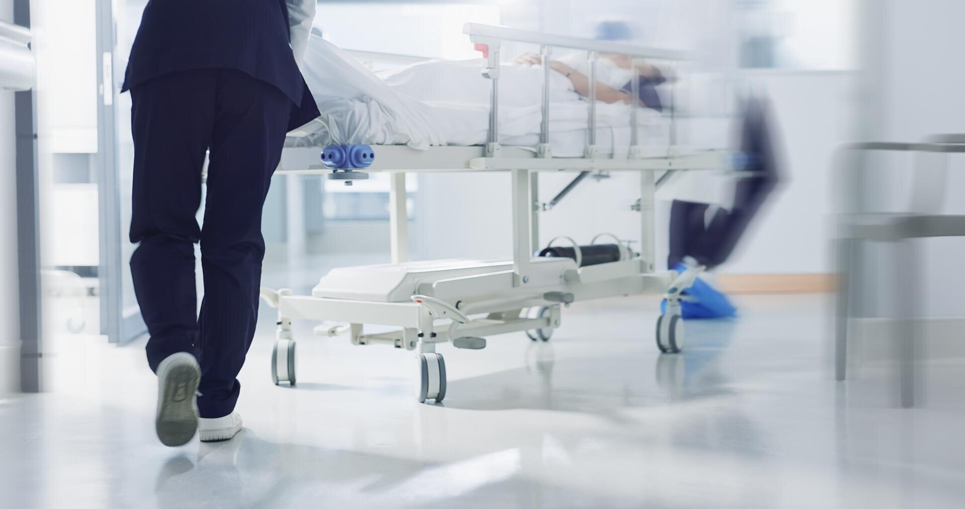 A medical professional pushes a hospital bed with a lying patient through a brightly lit hospital corridor. Another person in medical clothing can be seen in the background.