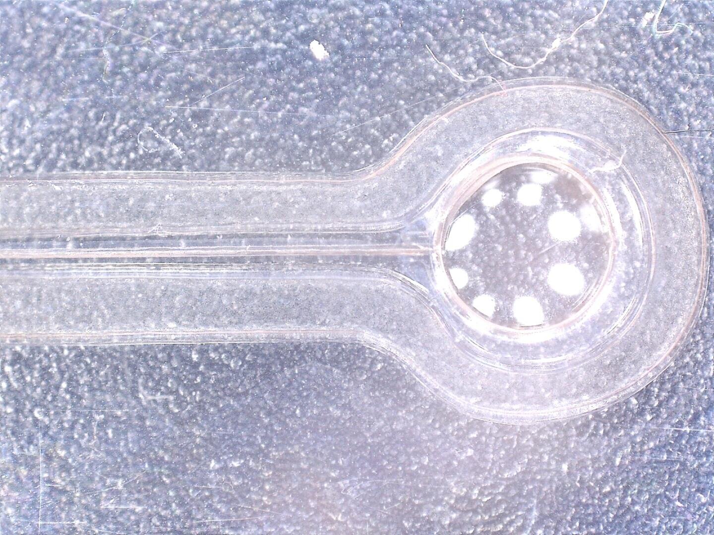 Nahaufnahme eines transparenten mikrofluidischen Geräts mit einer kreisförmigen Kammer, die fünf kleine weiße Punkte enthält, vor einem strukturierten, hellen Hintergrund.