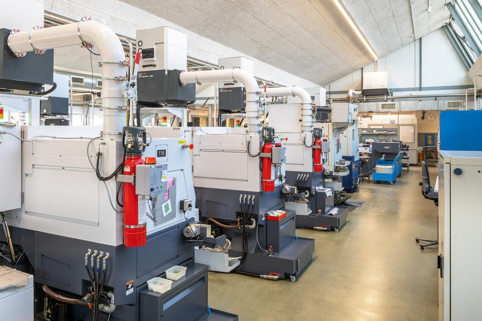 Eine Reihe moderner CNC-Maschinen mit weißen Lüftungsrohren und angebrachten Feuerlöschern säumen eine saubere, helle Industriehalle. Der Raum hat hohe Decken, große Fenster und übersichtlich angeordnete Arbeitsplätze.