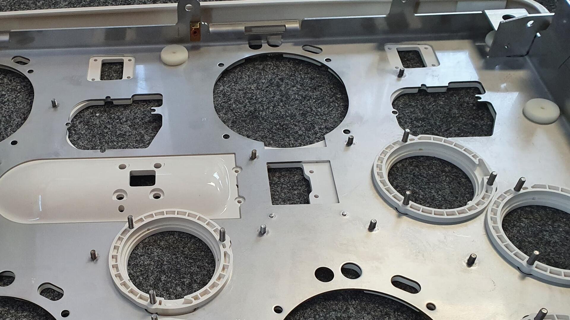 Close-up of a metal plate with circular and rectangular cutouts, white plastic rings and several metal posts resting on a textured gray surface, possibly part of an appliance or machine assembly.