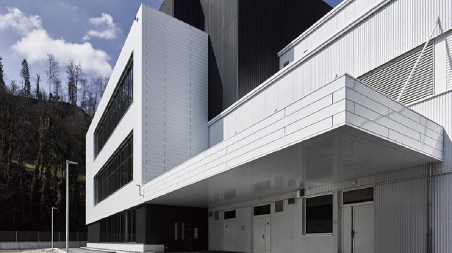 Ein modernes weißes Gebäude mit scharfen Winkeln und großen Fenstern, mit Metallverkleidung und mehreren Stockwerken, steht unter einem teilweise bewölkten Himmel mit sichtbaren Bäumen im Hintergrund.