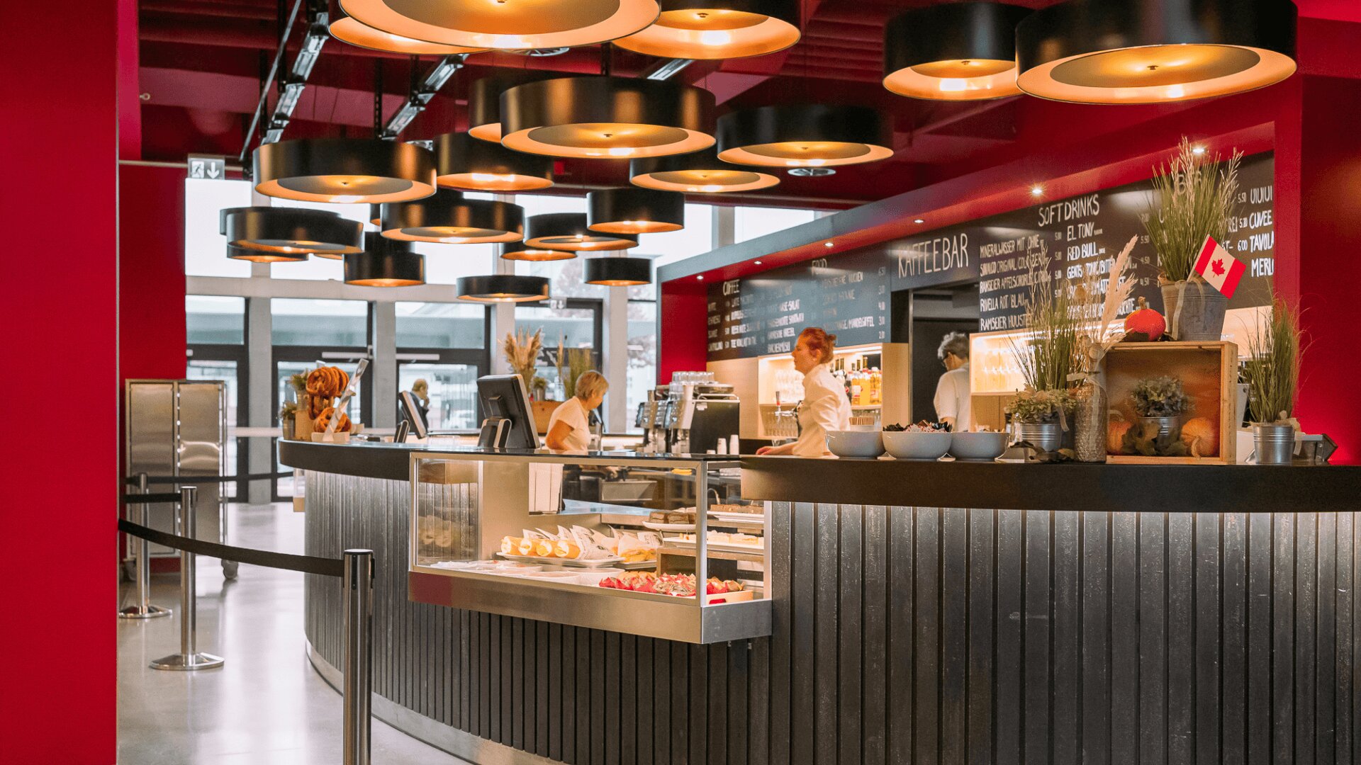 Ein modernes Café mit roten Wänden, runden Hängelampen und einer geschwungenen Theke mit Gebäck. Mitarbeiter in weißen Uniformen bereiten Bestellungen vor, und hinter der Theke stehen Regale mit Pflanzen. Im Hintergrund ist eine Speisekarte auf einer Kreidetafel zu sehen.
